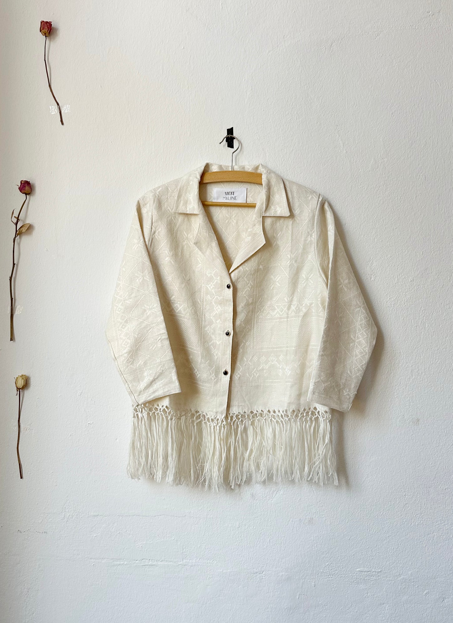 A cream blouse with fringed hem and vintage buttons hangs on a wall