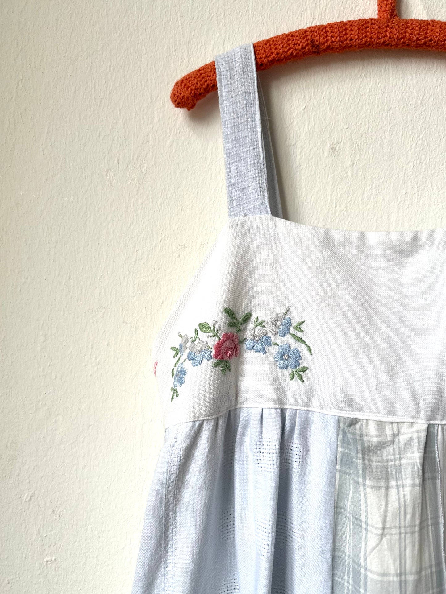 A detail view of the embroidery on the op front of a white and blue dress hanging on a wall.