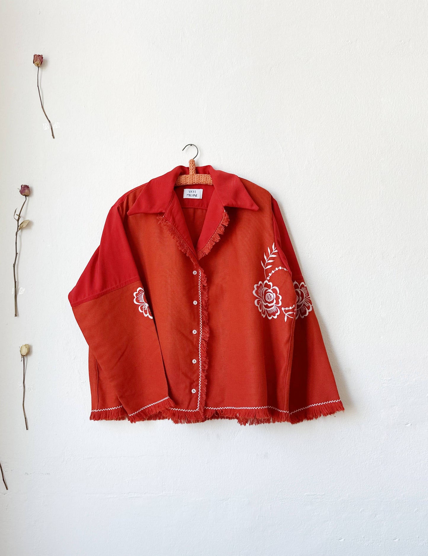A red and white embroidered shirt with fringe trim hangs on a wall next to some dried roses.