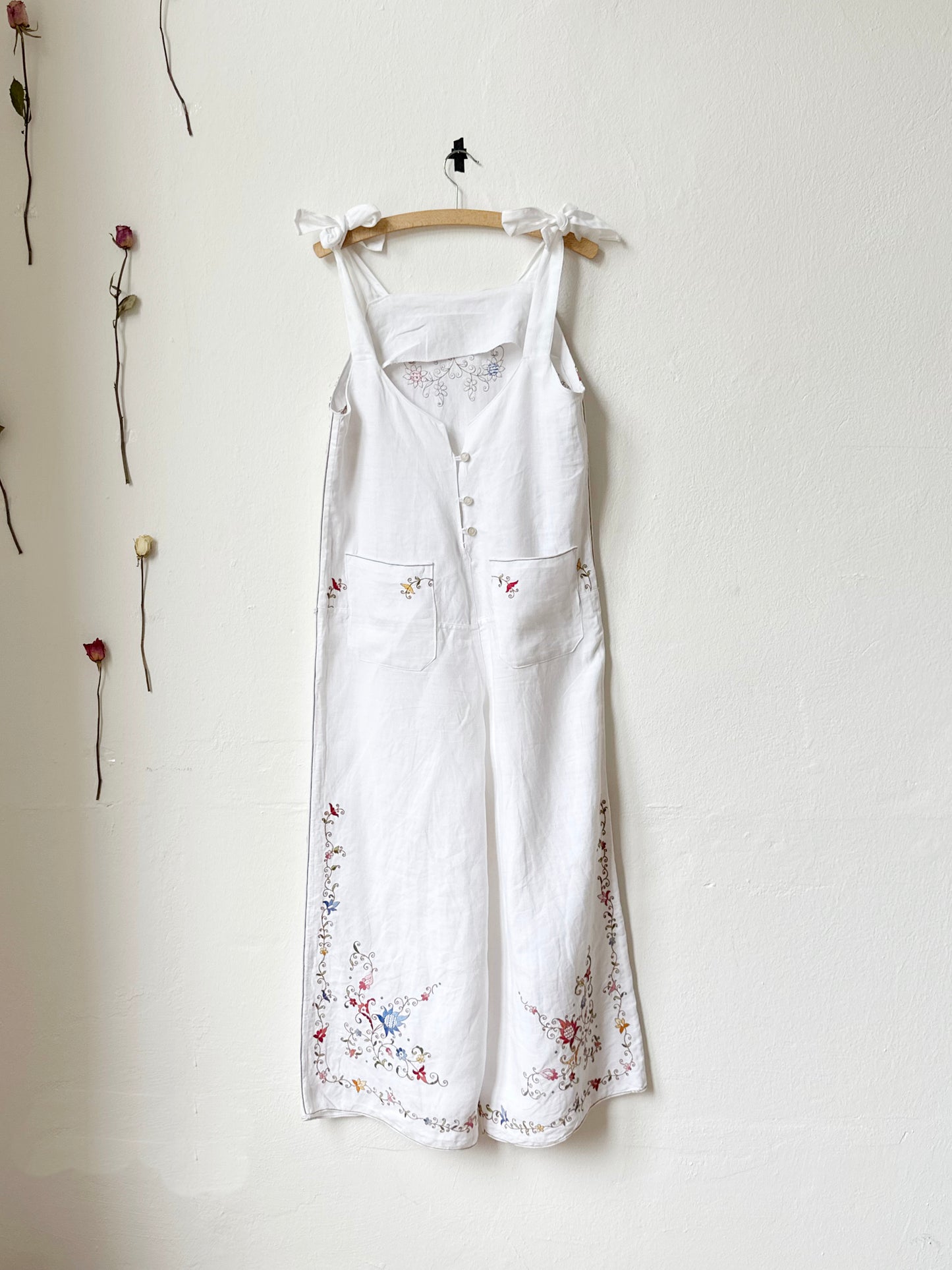 Back view of White overalls/jumpsuit with colourful floral embroidery hanging on a wall.