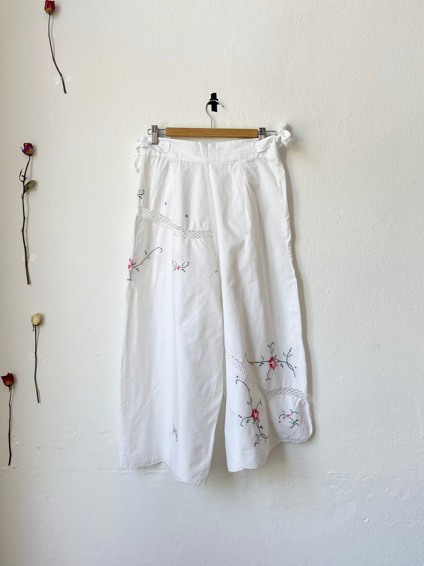 White embroidered wide leg trousers with side ties hang on the wall with the back facing the camera.