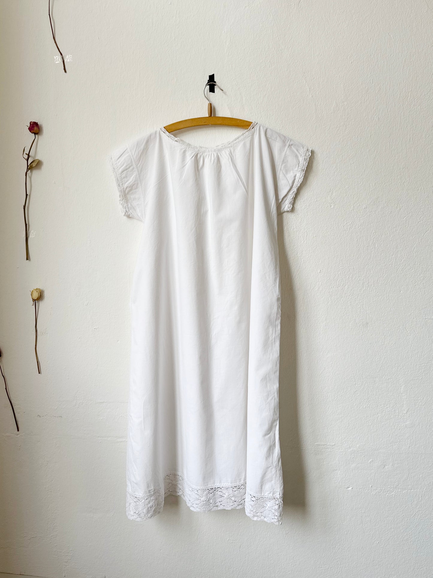 White dress hanging on a wooden hanger against a white wall with a single red flower to the left.