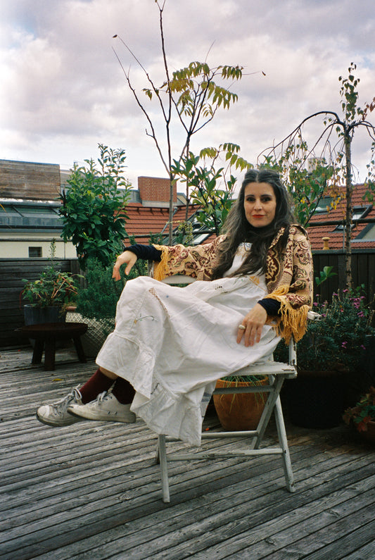 vicki sits sideways in a chair on a terrace wearing a dress and cropped blanket jacket