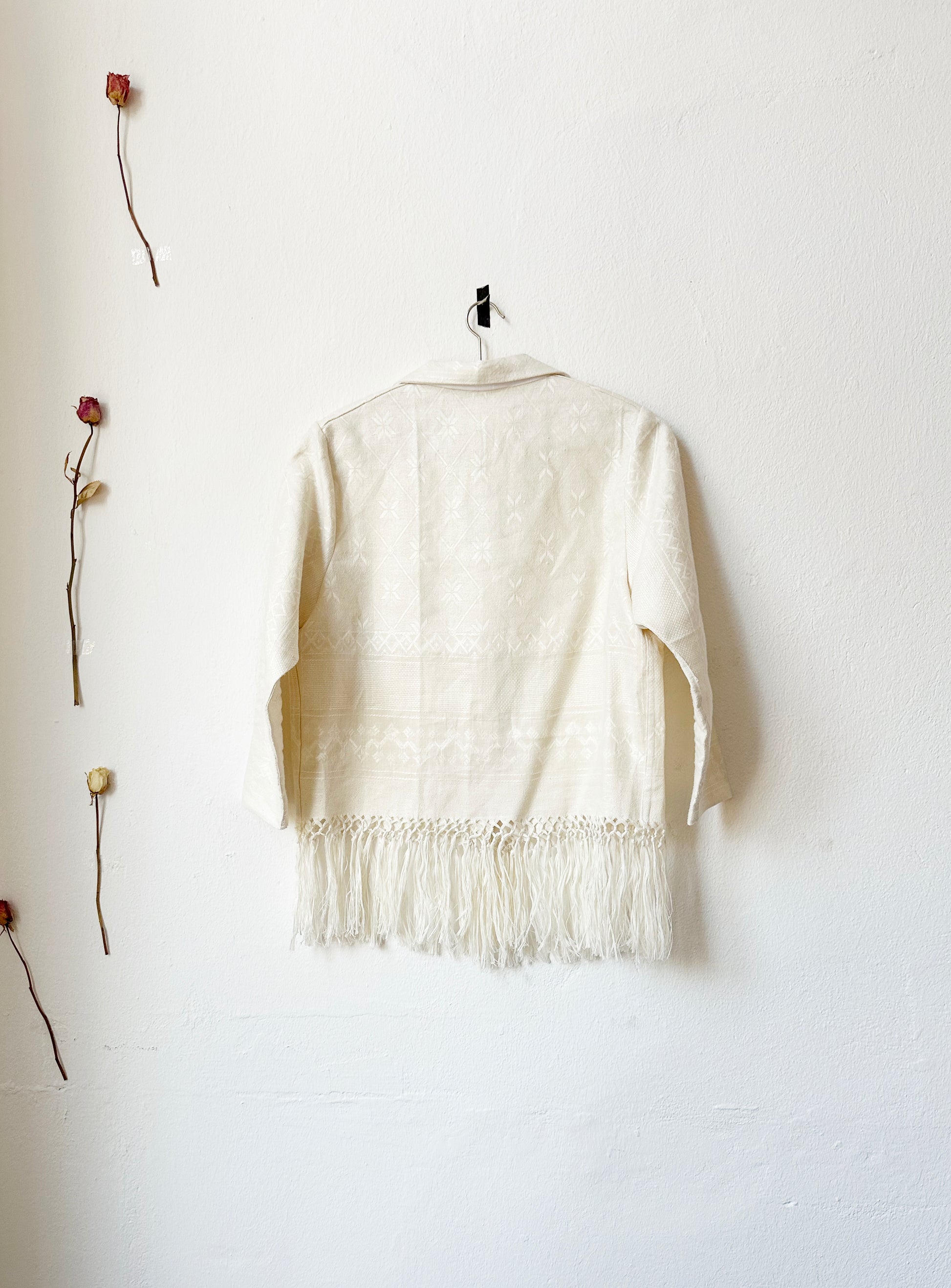 A blouse with fringe hem hangs on the wall with the back facing viewer