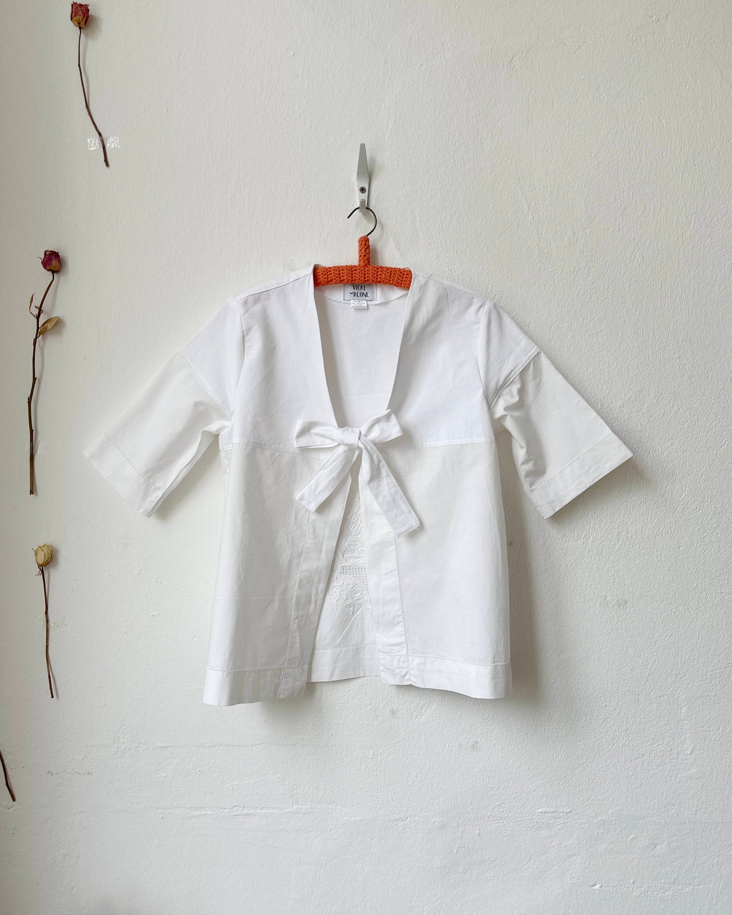 White blouse with a bow tie on a hanger against a plain wall.