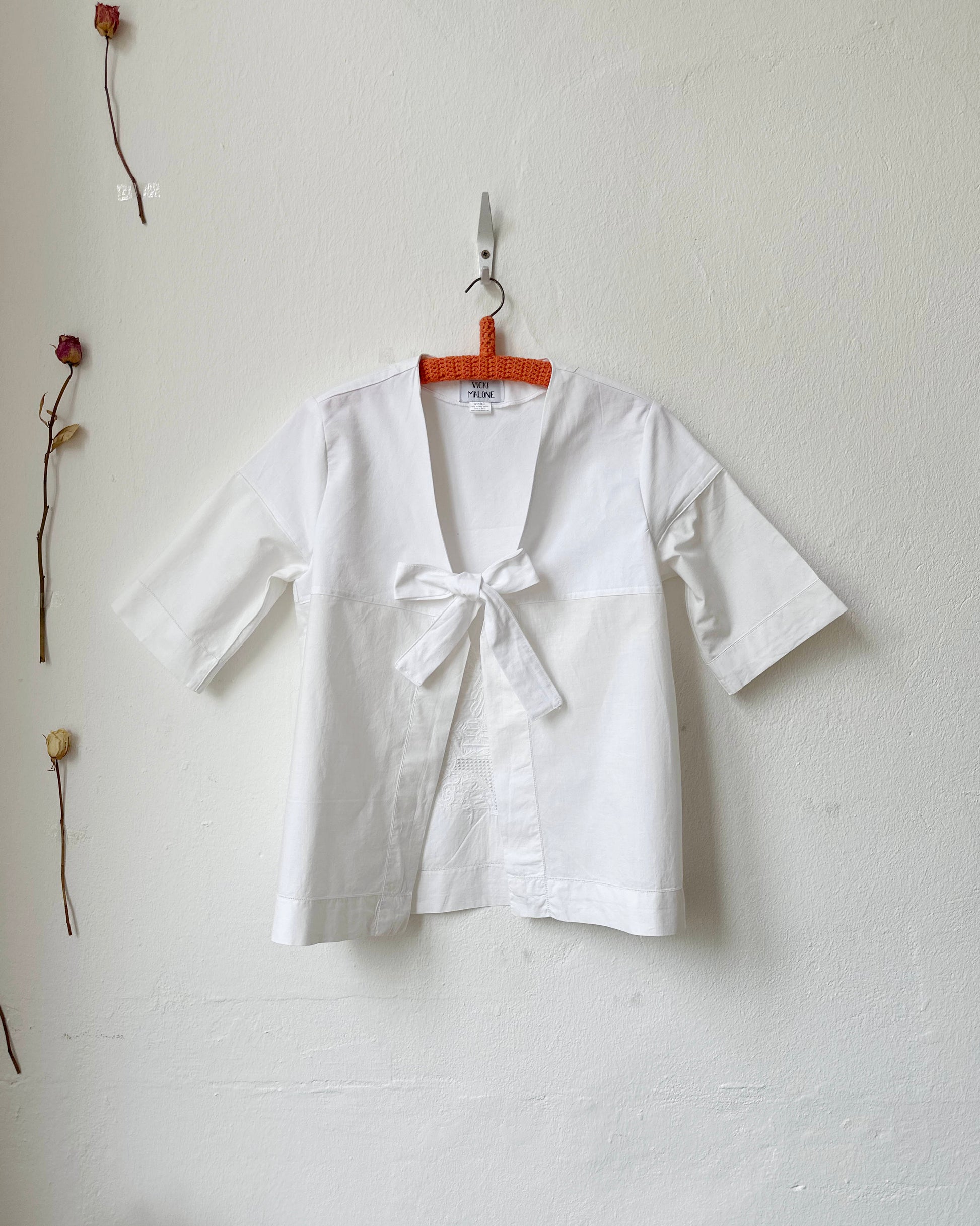 White blouse with a bow tie on a hanger against a plain wall.