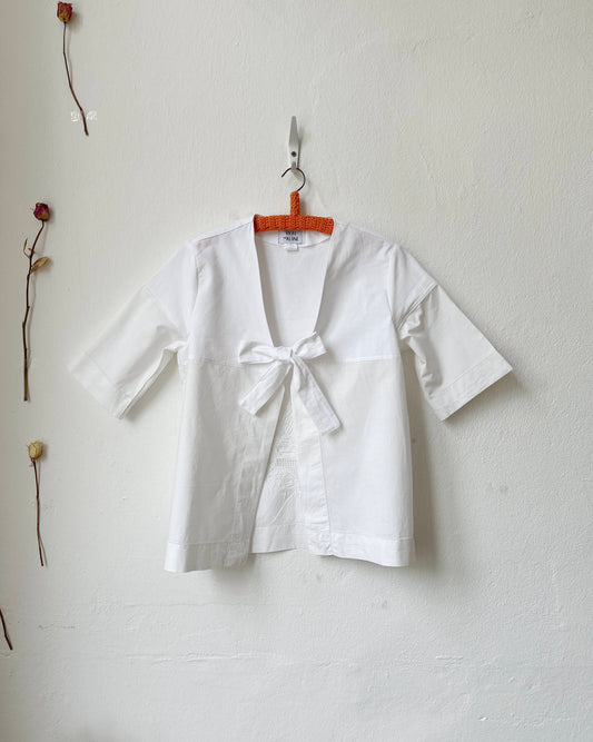 White blouse with a bow tie on a hanger against a plain wall.