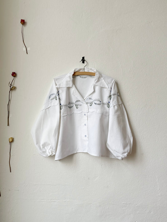 A white blouse with buttoned front fastening and elasticated cuff sleeves, displayed on a hanger against a plain wall. The blouse appears to be made from a vintage textile with a Christmas candle pattern.