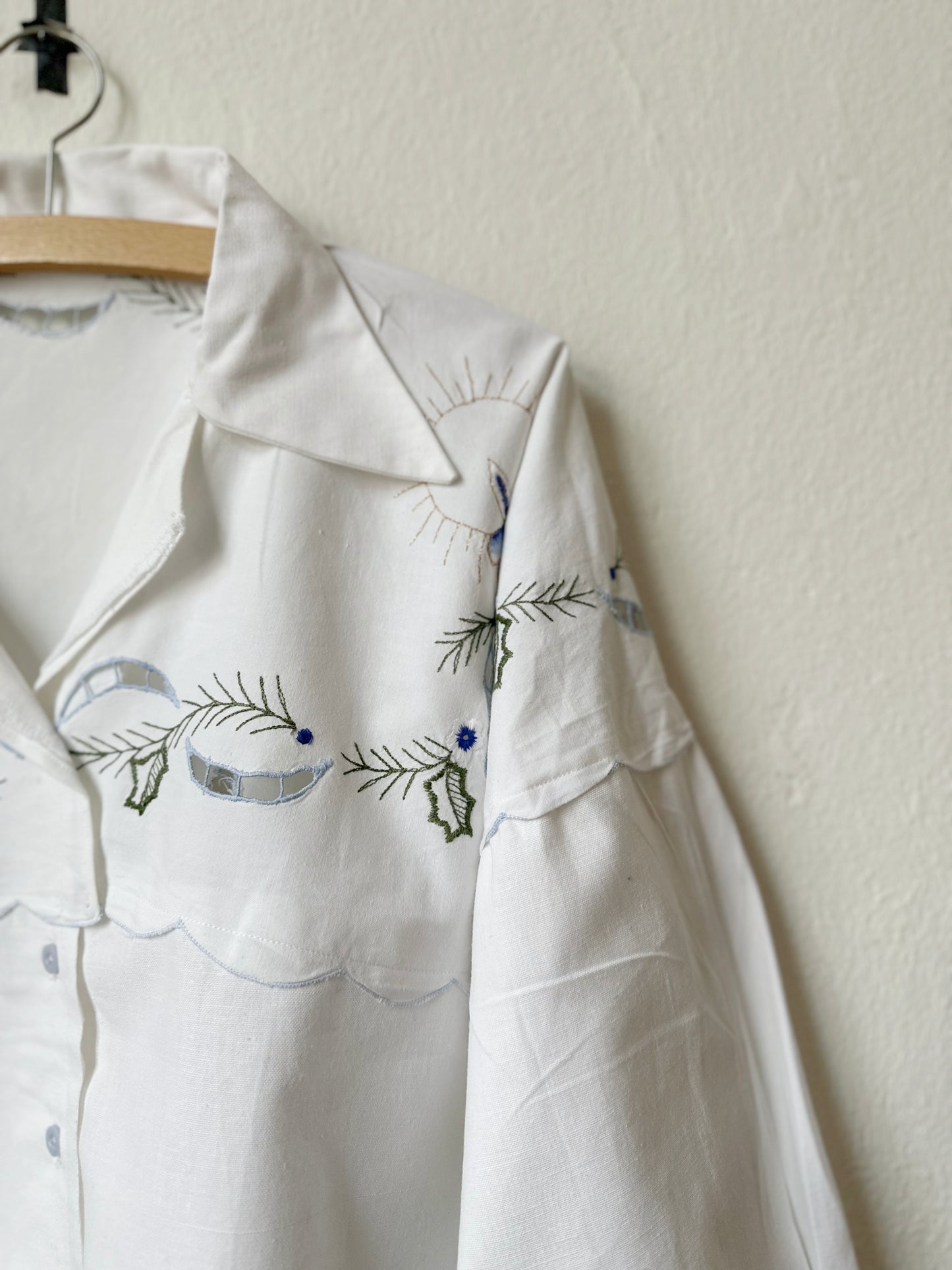 A detail view of the embroidery of a white blouse hanging on a wall.