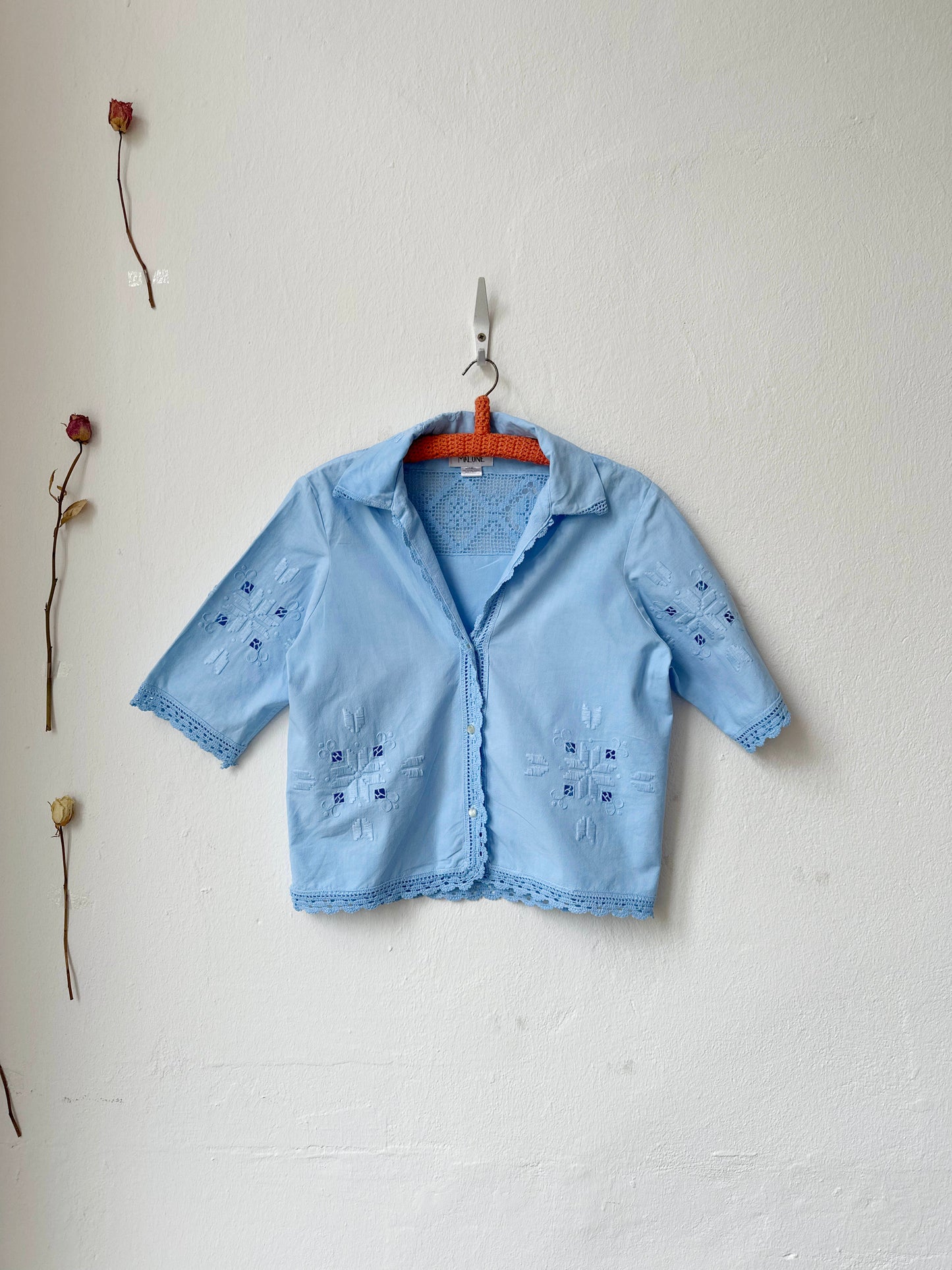 Light blue embroidered shirt on a hanger against a white wall with dried flowers.