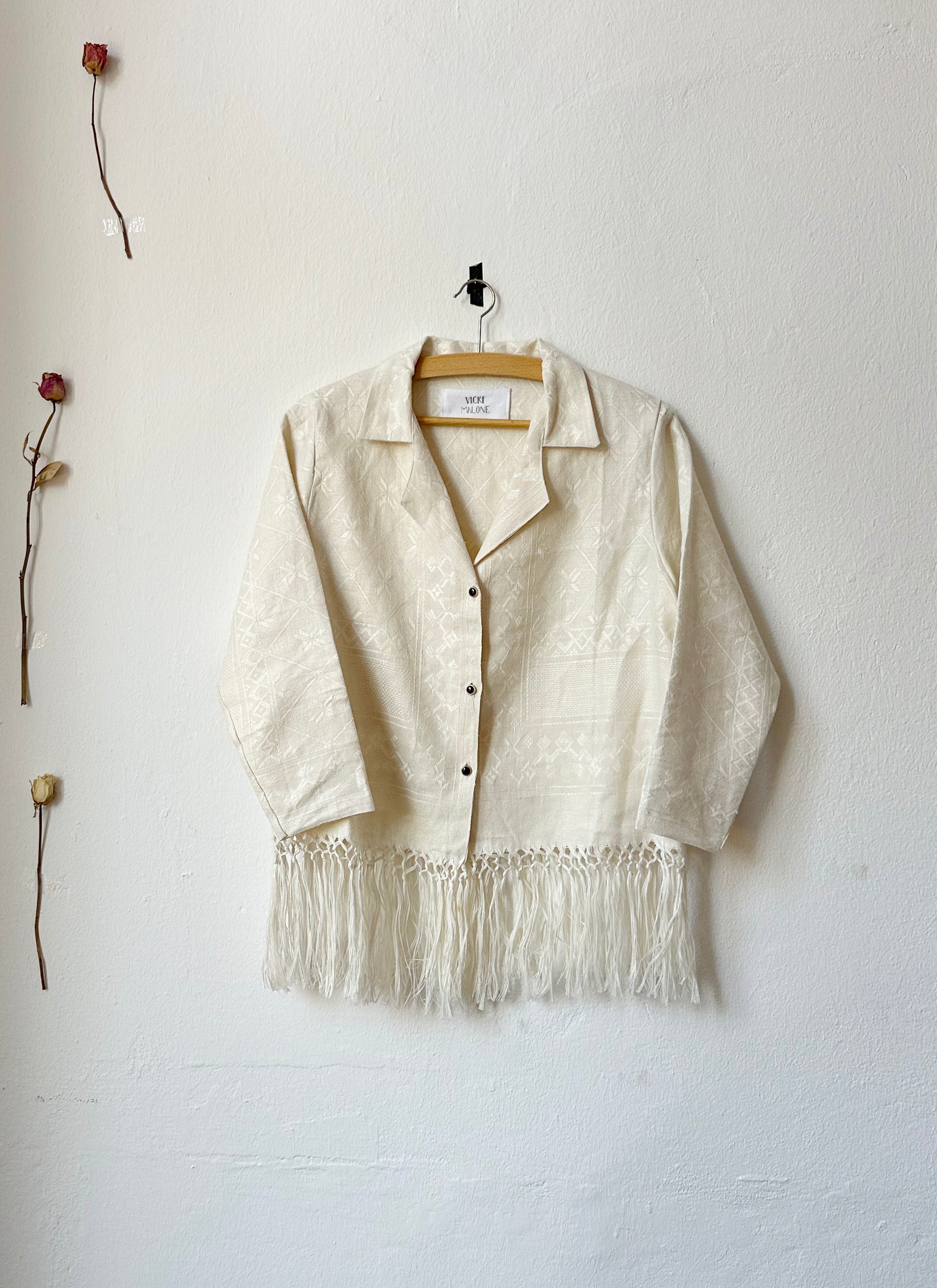 A cream blouse with fringed hem and vintage buttons hangs on a wall