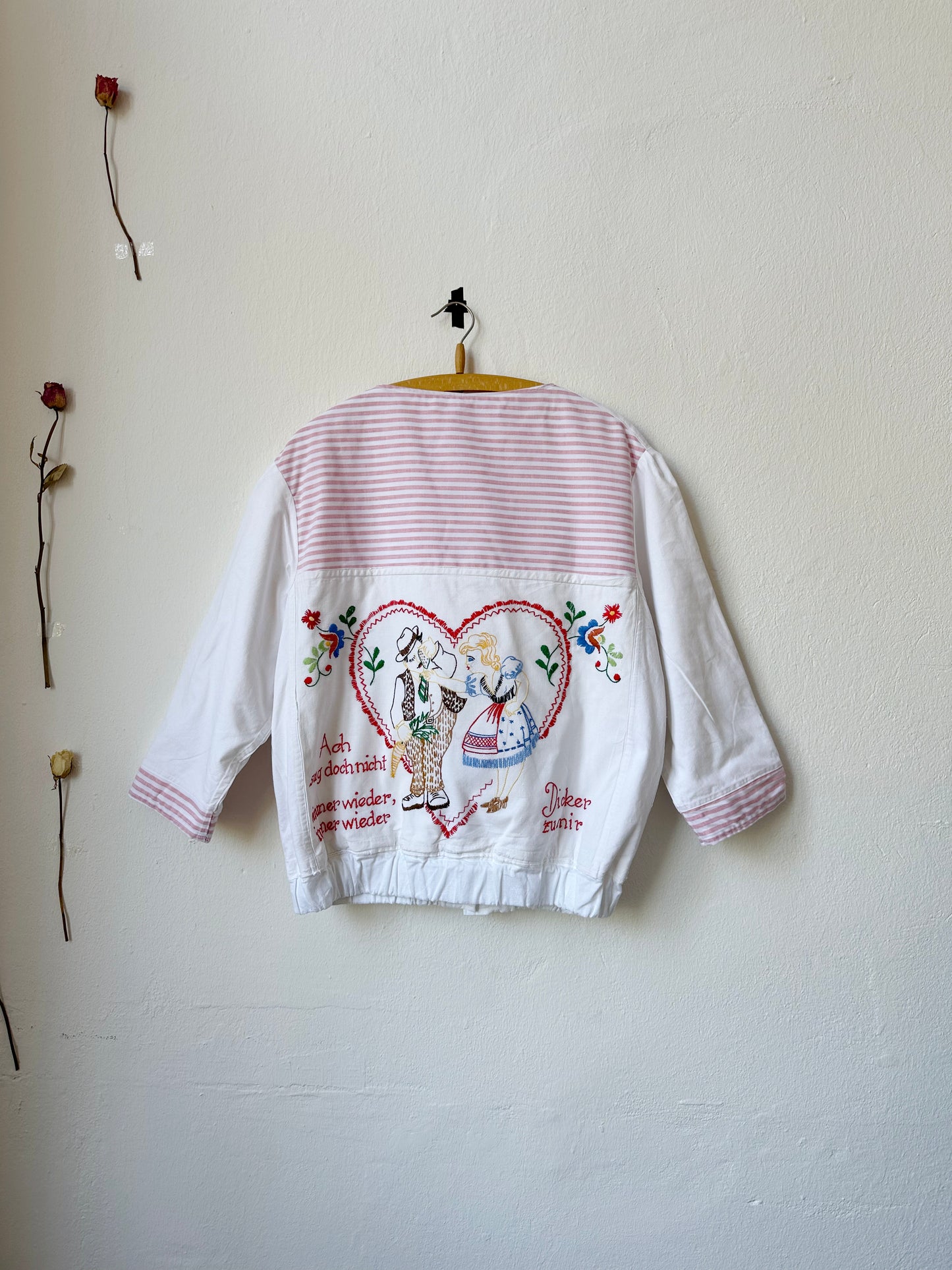 A patchwork chore jacket with embroidered german phrases and pictures hangs on a wall with the back facing out.
