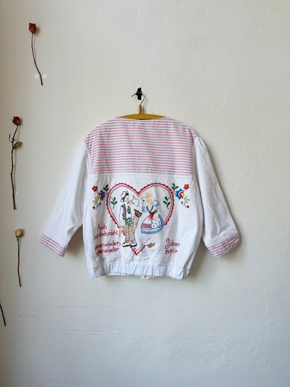 A patchwork chore jacket with embroidered german phrases and pictures hangs on a wall with the back facing out.
