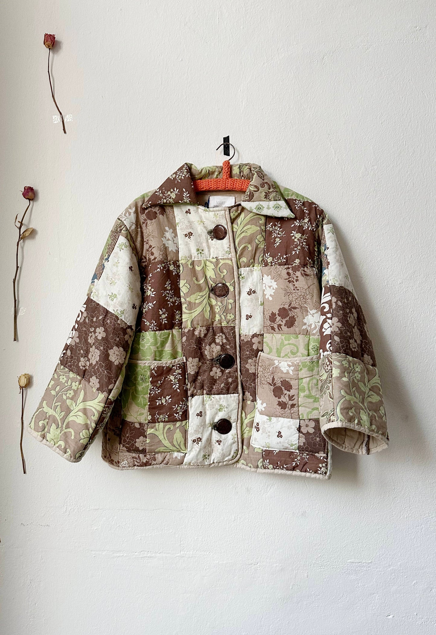 A multicolored quilted jacket with a geometric pattern, featuring a button front and pockets at the hip, displayed on a hanger against a white wall with a few dried flowers decoration.