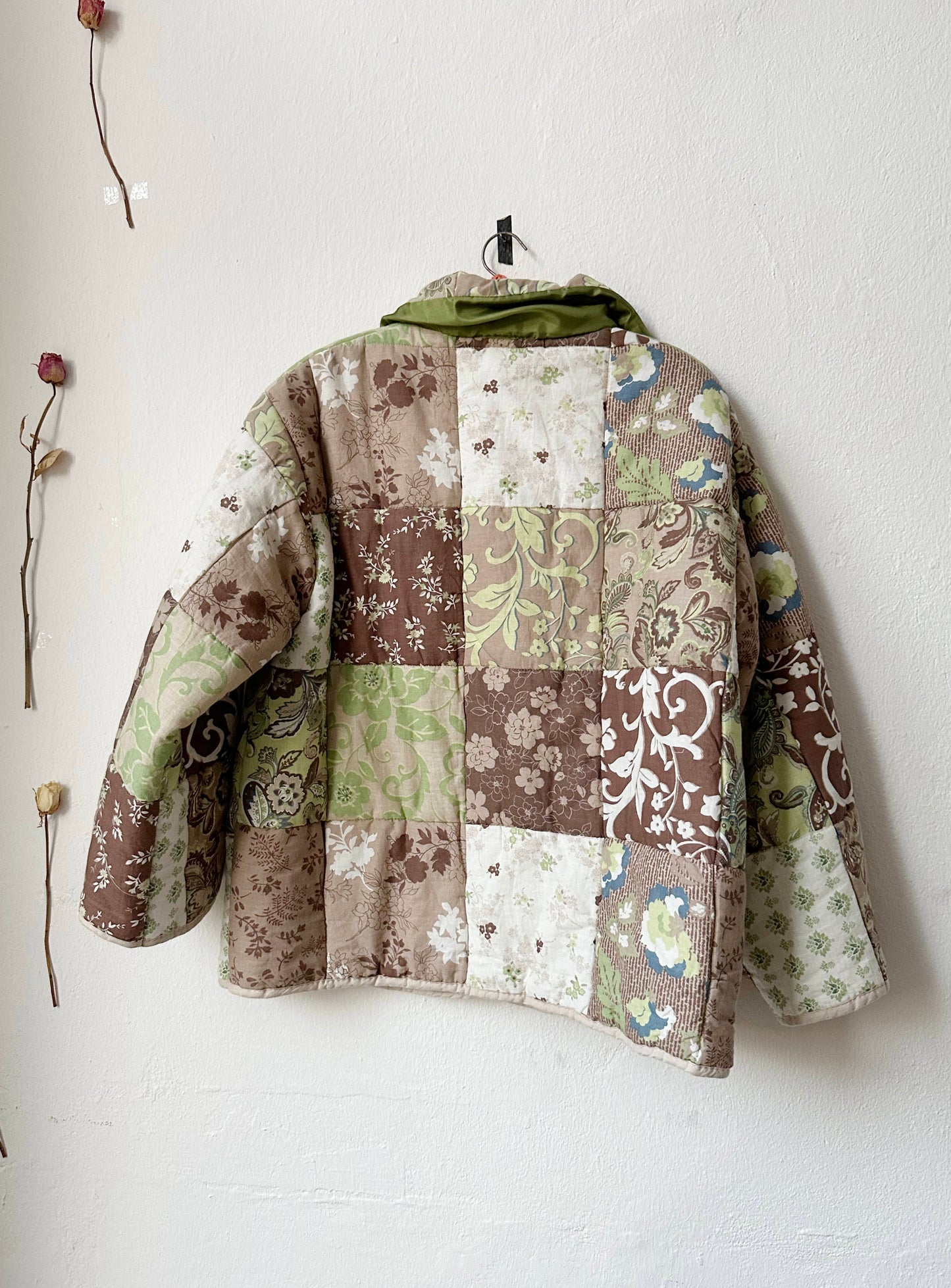 Back view of a brown and green patterned quilt jacket on a hanger against a white wall.