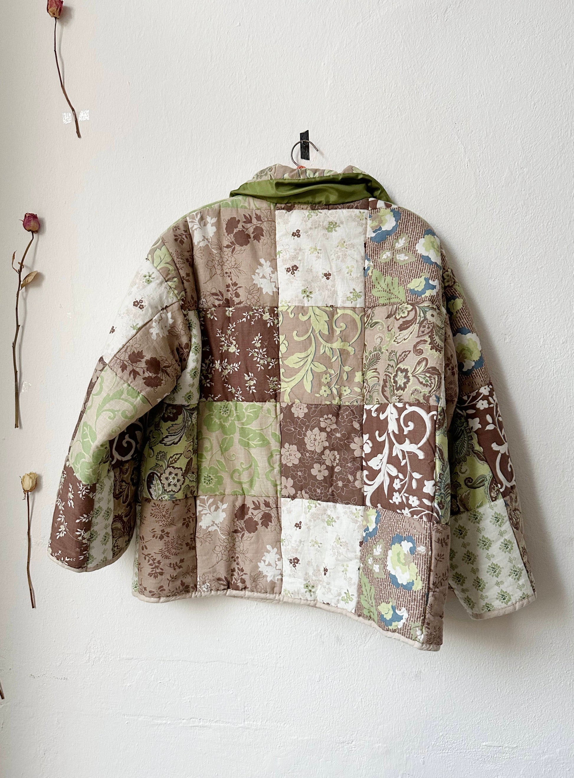 Back view of a brown and green patterned quilt jacket on a hanger against a white wall.