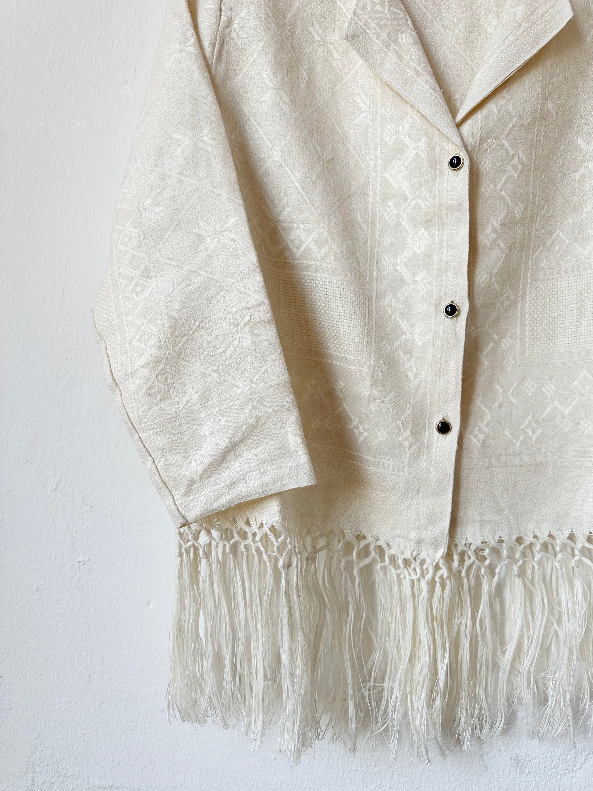 A close up view of the vintage buttons of a blouse with fringed hem hanging on a wall