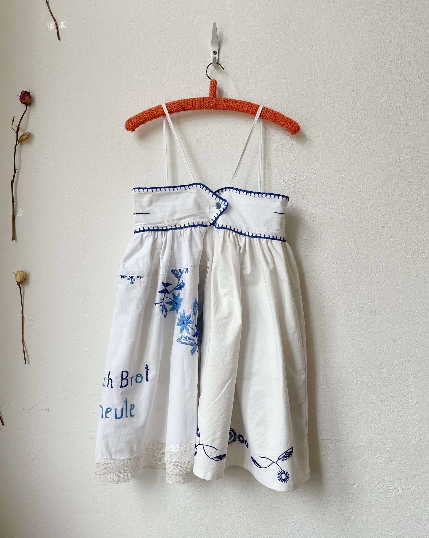 White dress with blue embroidery on a hanger against a white wall.