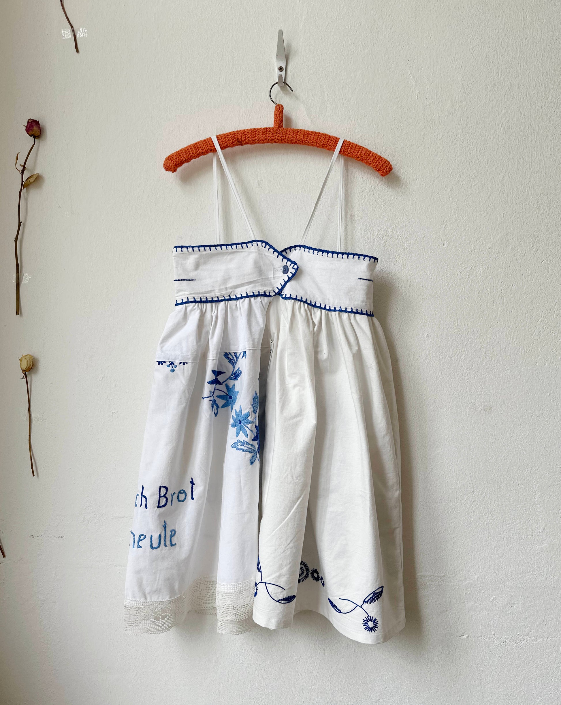 White dress with blue embroidery on a hanger against a white wall.