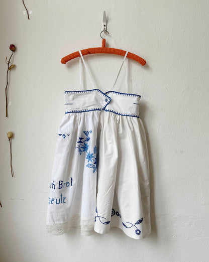White dress with blue embroidery on a hanger against a white wall.