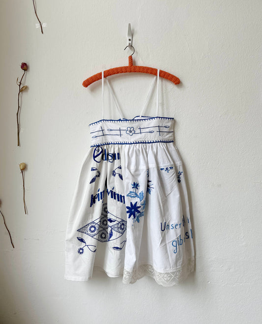 White dress with blue embroidery patterns hanging on a wooden hanger against a white wall.