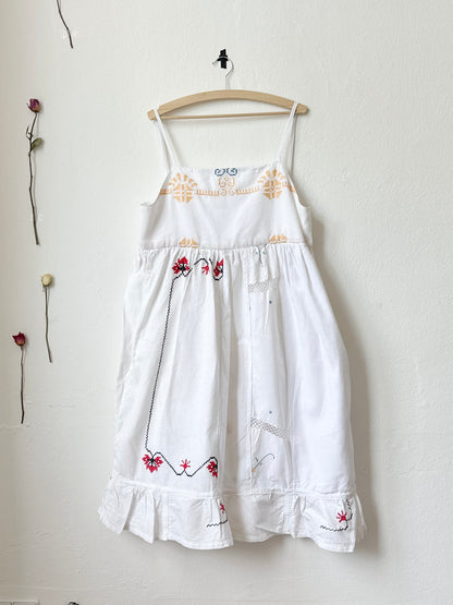 A white Ellen dress with patchwork details and pockets, displayed on a hanger against a plain wall, with dried flowers beside it.