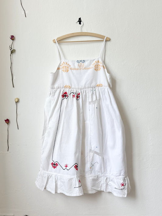 A white Ellen dress with patchwork details and pockets, displayed on a hanger against a plain wall, with dried flowers beside it.