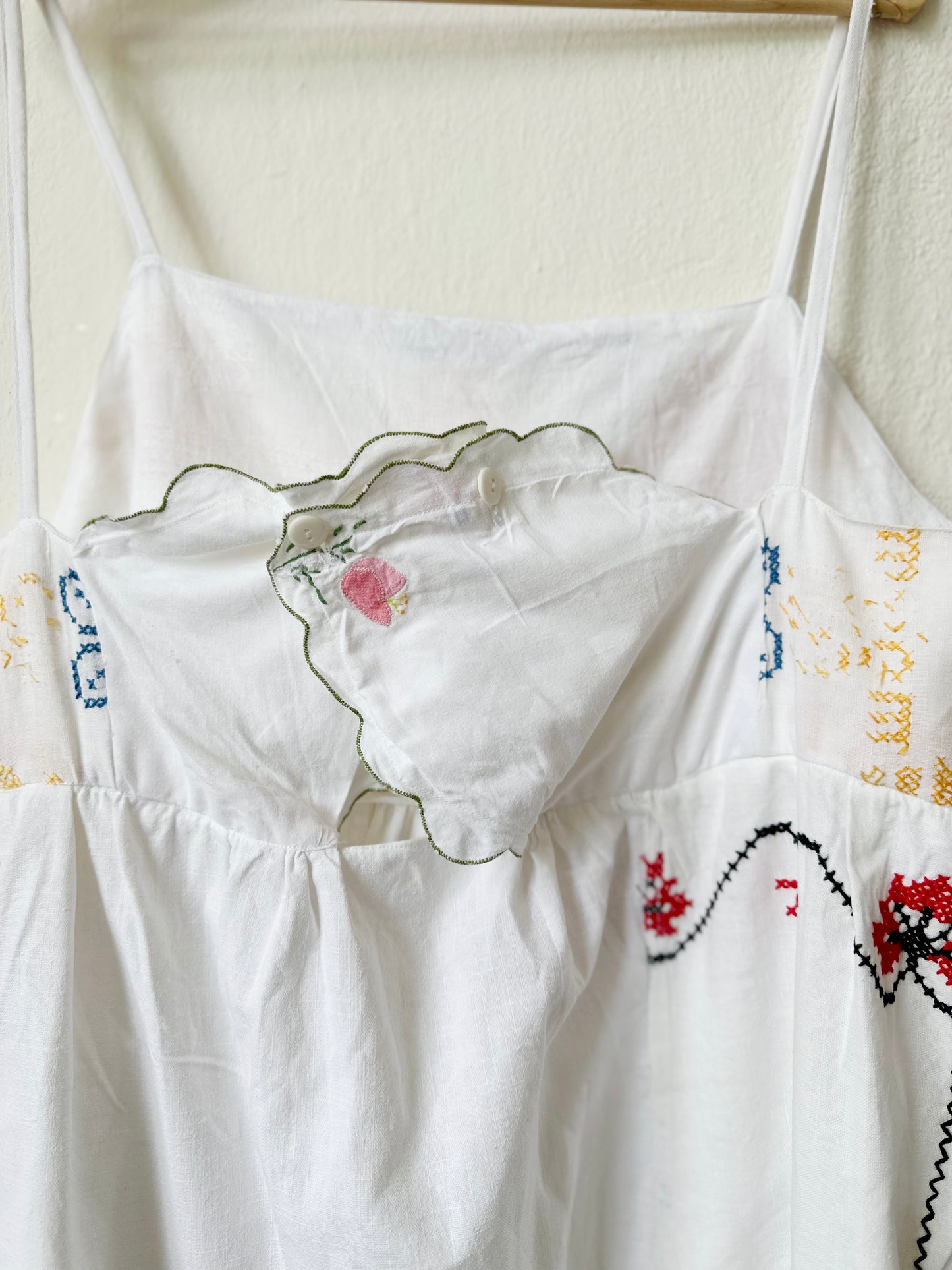 Back detail of a white Ellen dress with embroidered patchwork details displayed on a hanger against a plain wall.