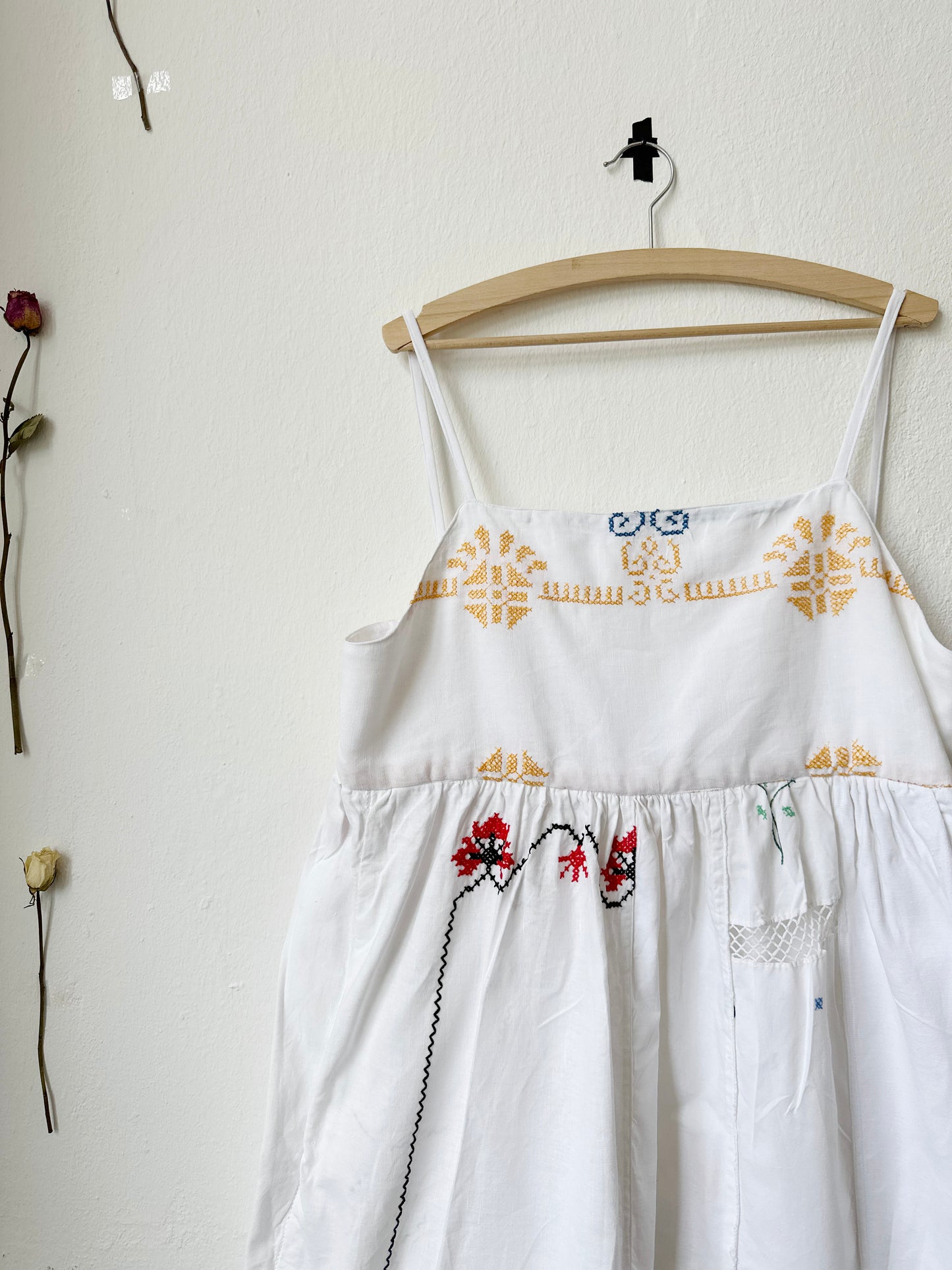 A white Ellen dress with patchwork details and pockets, displayed on a hanger against a plain wall, with dried flowers beside it.