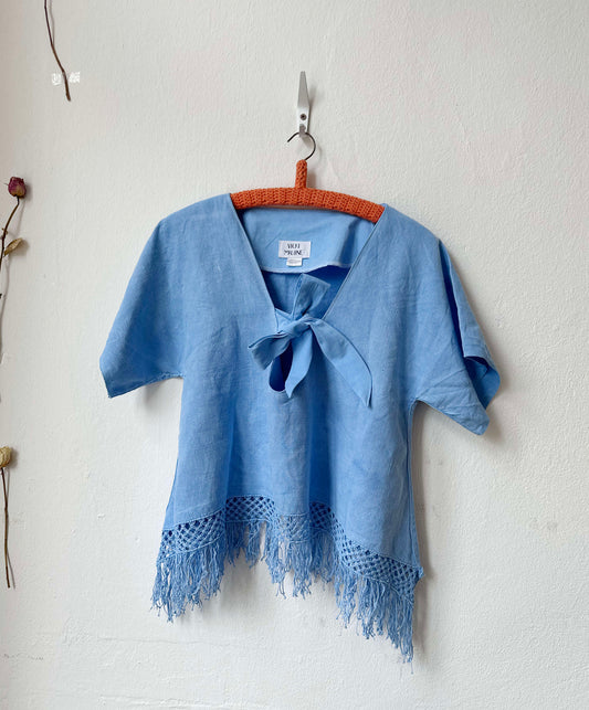 Blue top with fringe on a wooden hanger against a white wall.