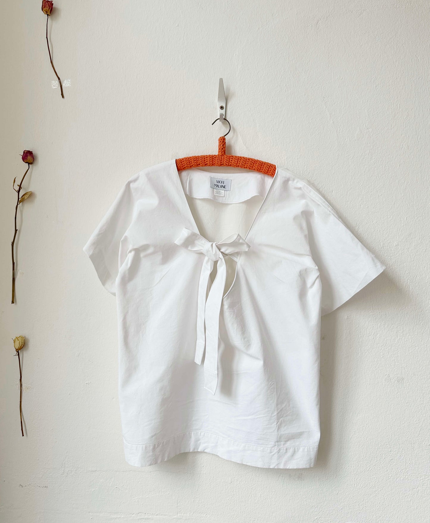 White blouse with a tie neckline hanging on a wooden hanger against a white wall.