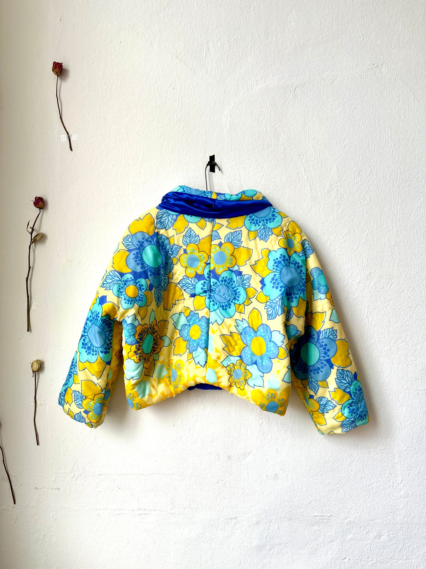 A yellow and blue floral DDR duvet jacket hangs on a white wall with the back facing out.