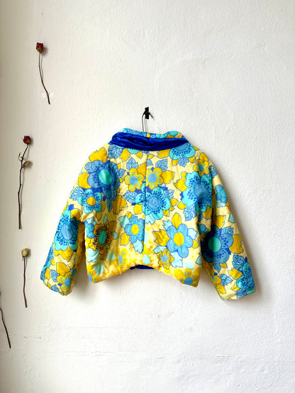 A yellow and blue floral DDR duvet jacket hangs on a white wall with the back facing out.