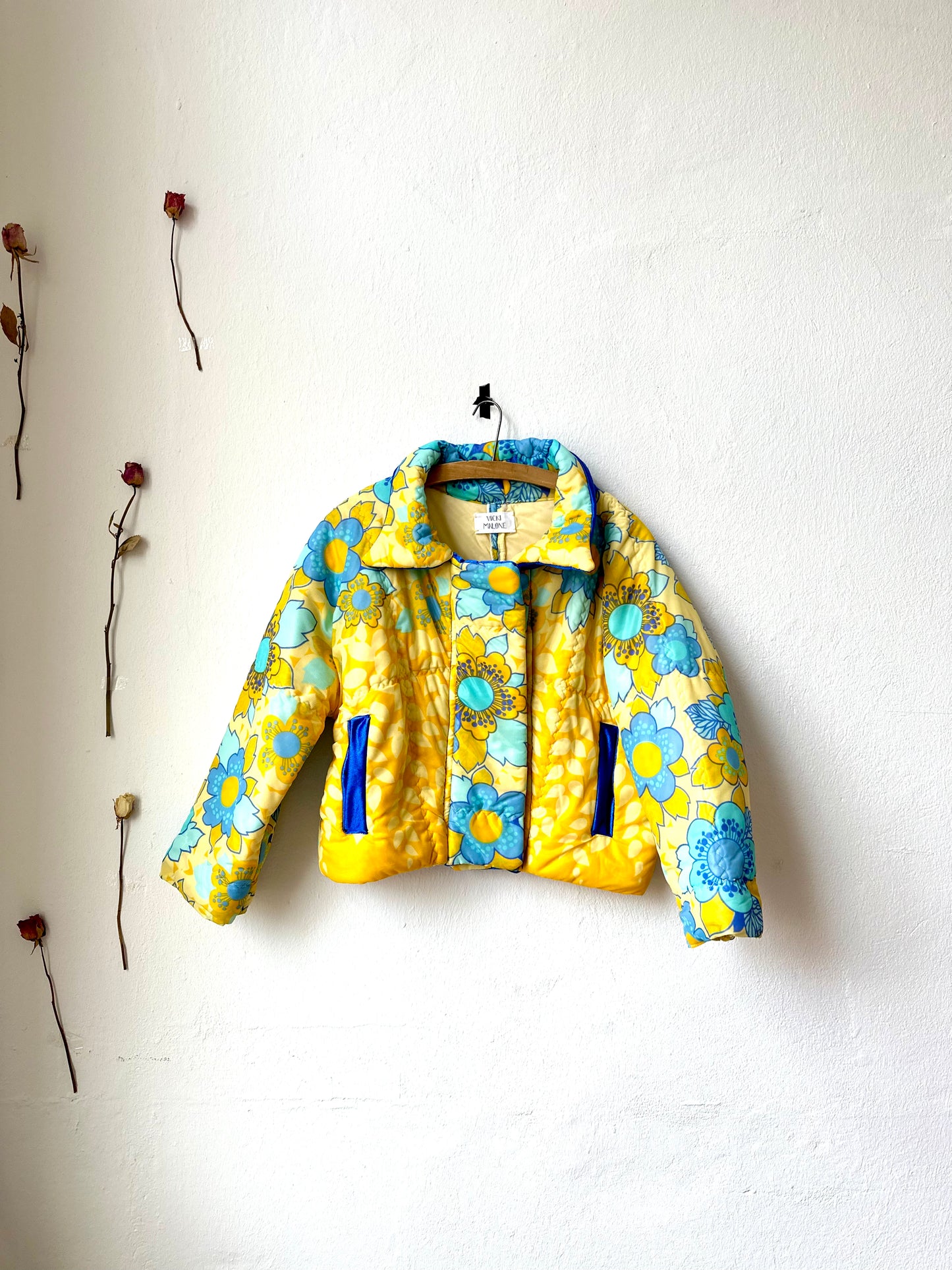 A vintage blue and yellow floral quilt jacket with wide neckline and snap fasteners, displayed on a hanger against a plain wall.