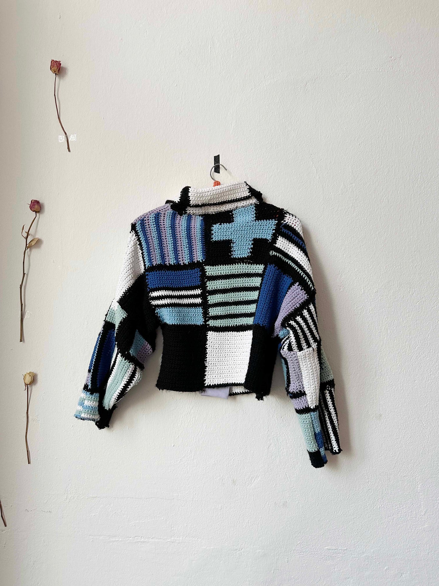 A crochet blanket jacket hangs on a wall with the back facing us. 