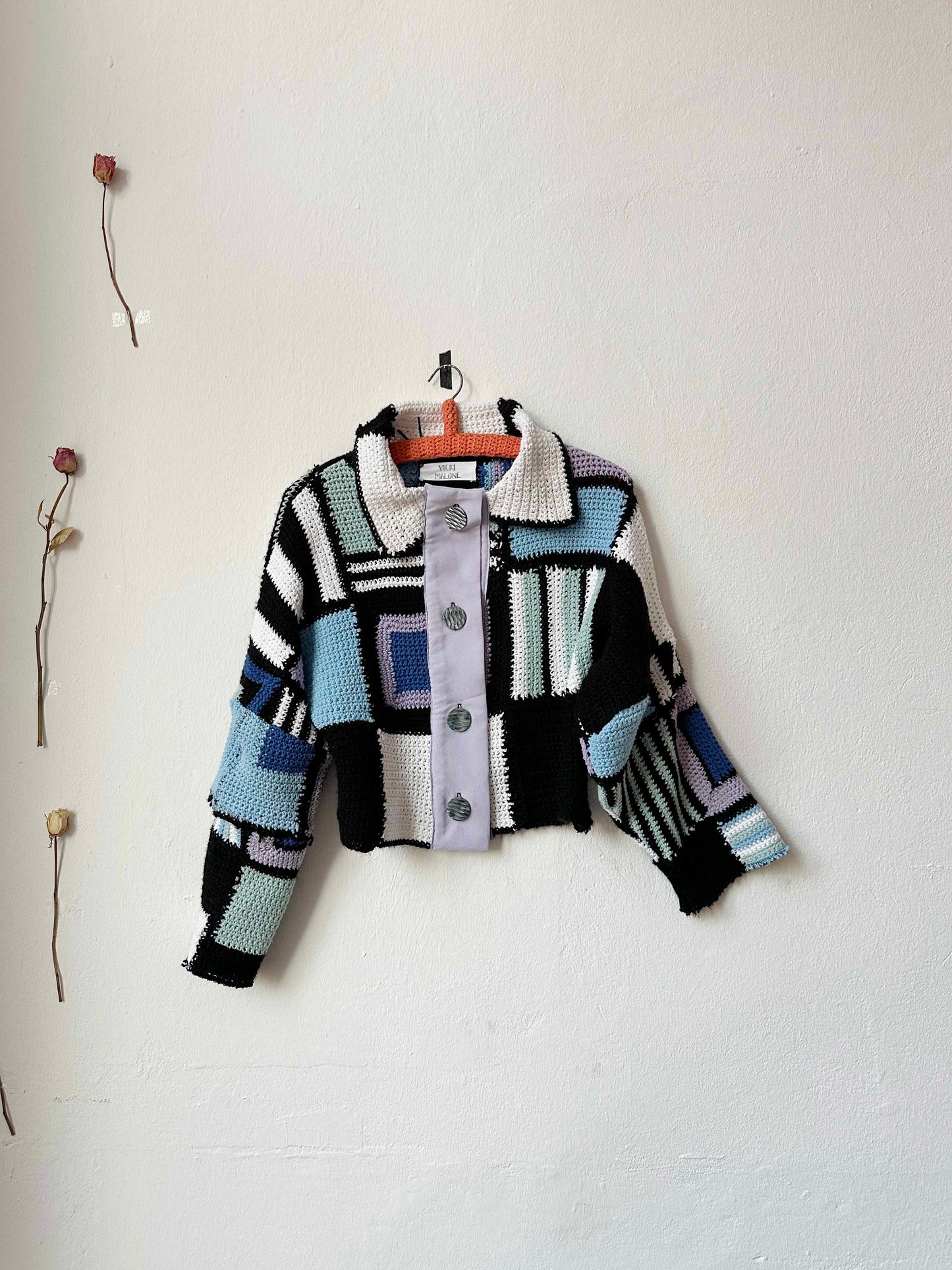 A crocheted blanket jacket in black, blue, and multicolor stripes, with a lilac front placket and mock mother of pearl buttons, displayed on a white wall.