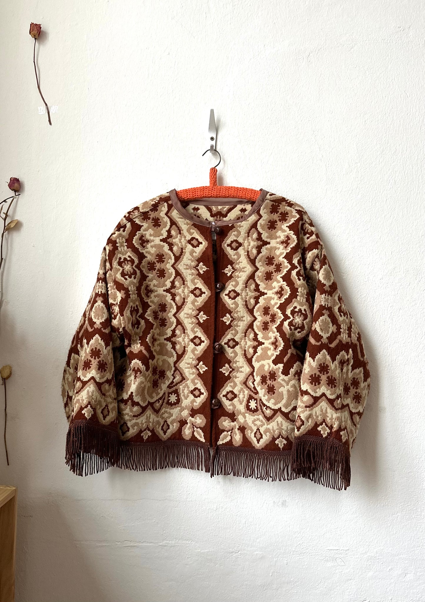 Knitted patterned jacket with fringes on a white wall.
