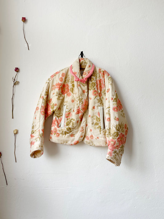 A floral duvet jacket with snap fasteners and a tie at the neck, displayed against a white background, with dried flowers arranged around it.