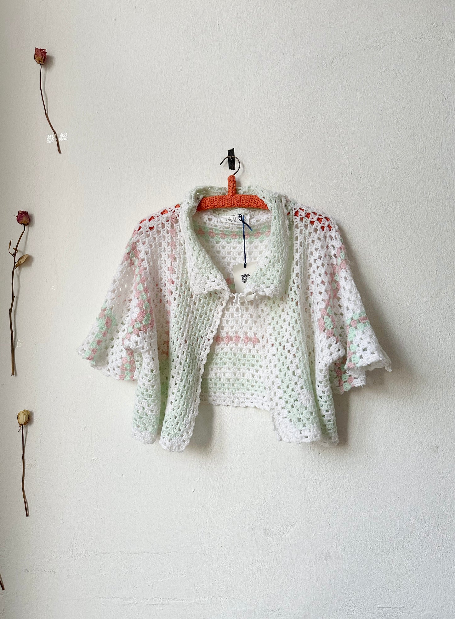 A crochet blanket jacket with a tie at the neck on a hanger against a white wall.