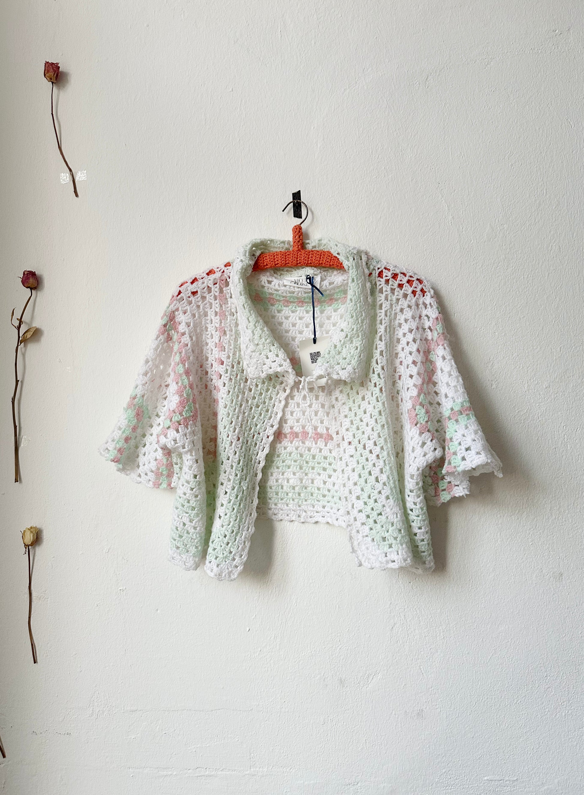 A crochet blanket jacket with a tie at the neck on a hanger against a white wall.