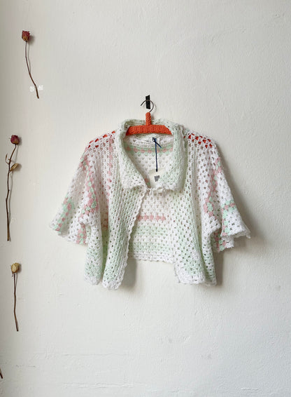A crochet blanket jacket with a tie at the neck on a hanger against a white wall.