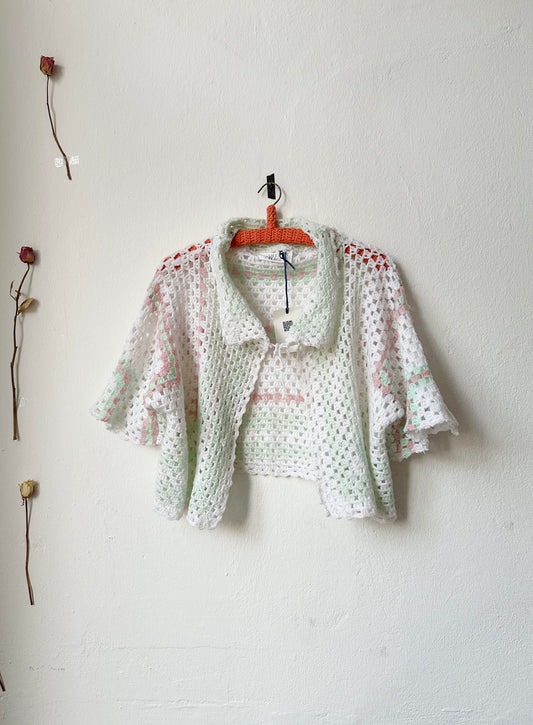 A crochet blanket jacket with a tie at the neck on a hanger against a white wall.