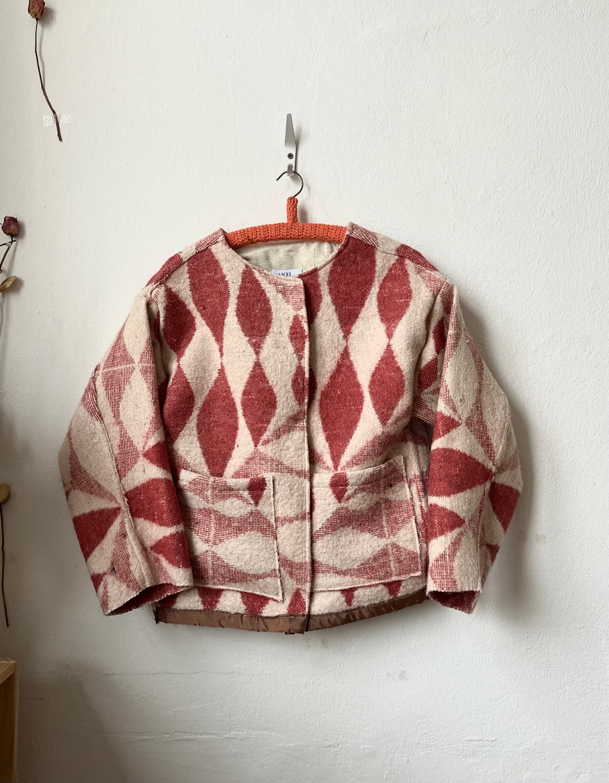Blanket jacket with red and beige geometric pattern hanging on a hook against a white wall.
