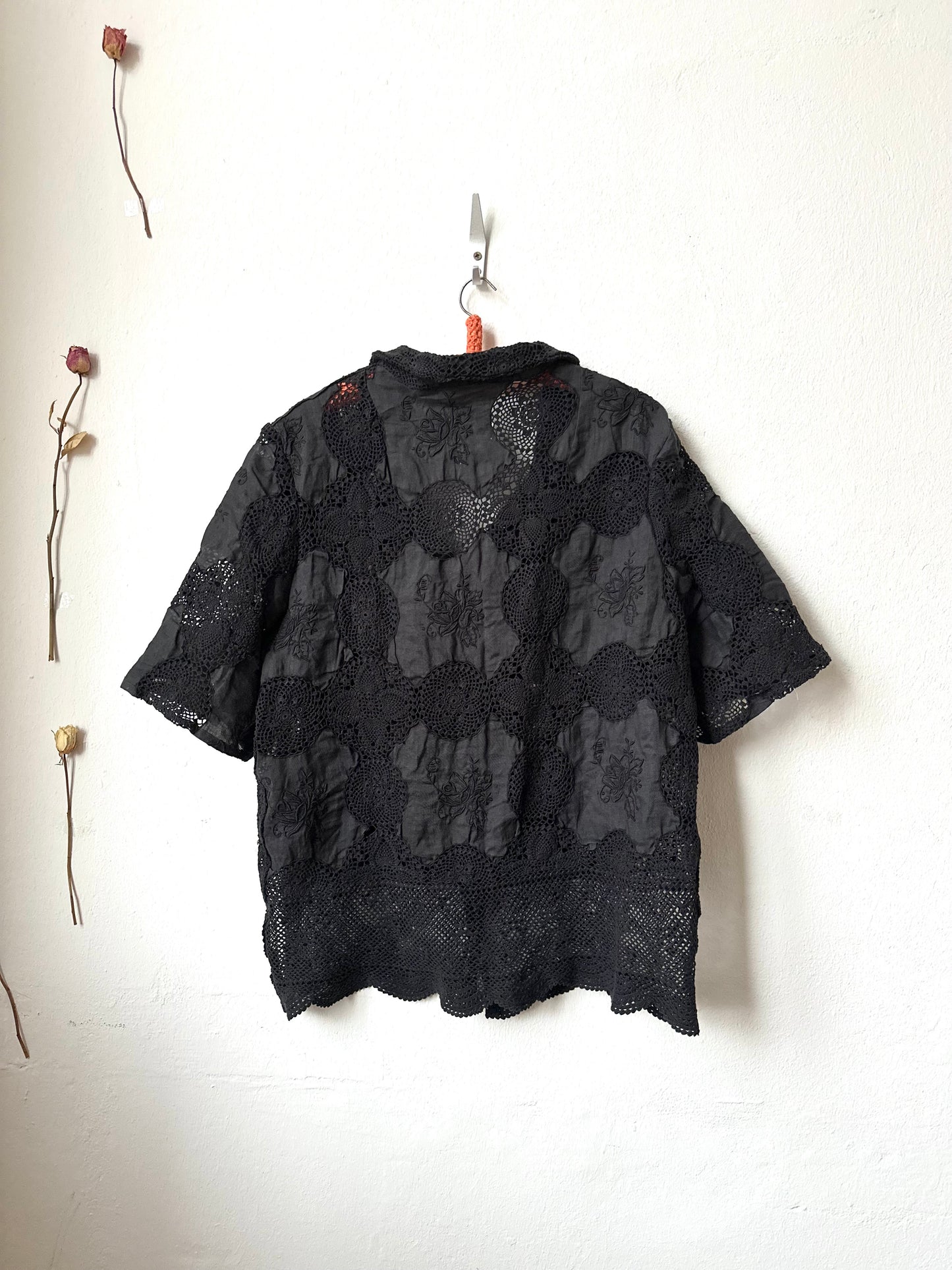 Black floral-patterned lace shirt hanging on a white wall with dried flowers.