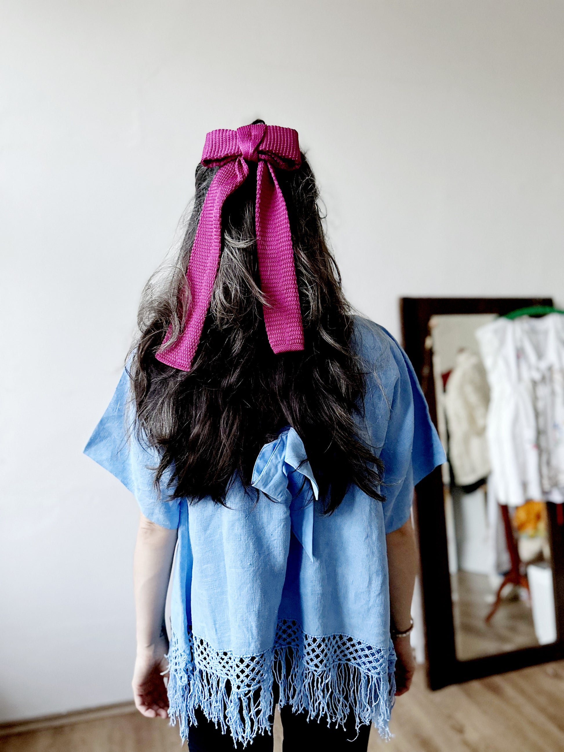 Person wearing a blue top with a pink knitted bow, standing in front of a mirror.