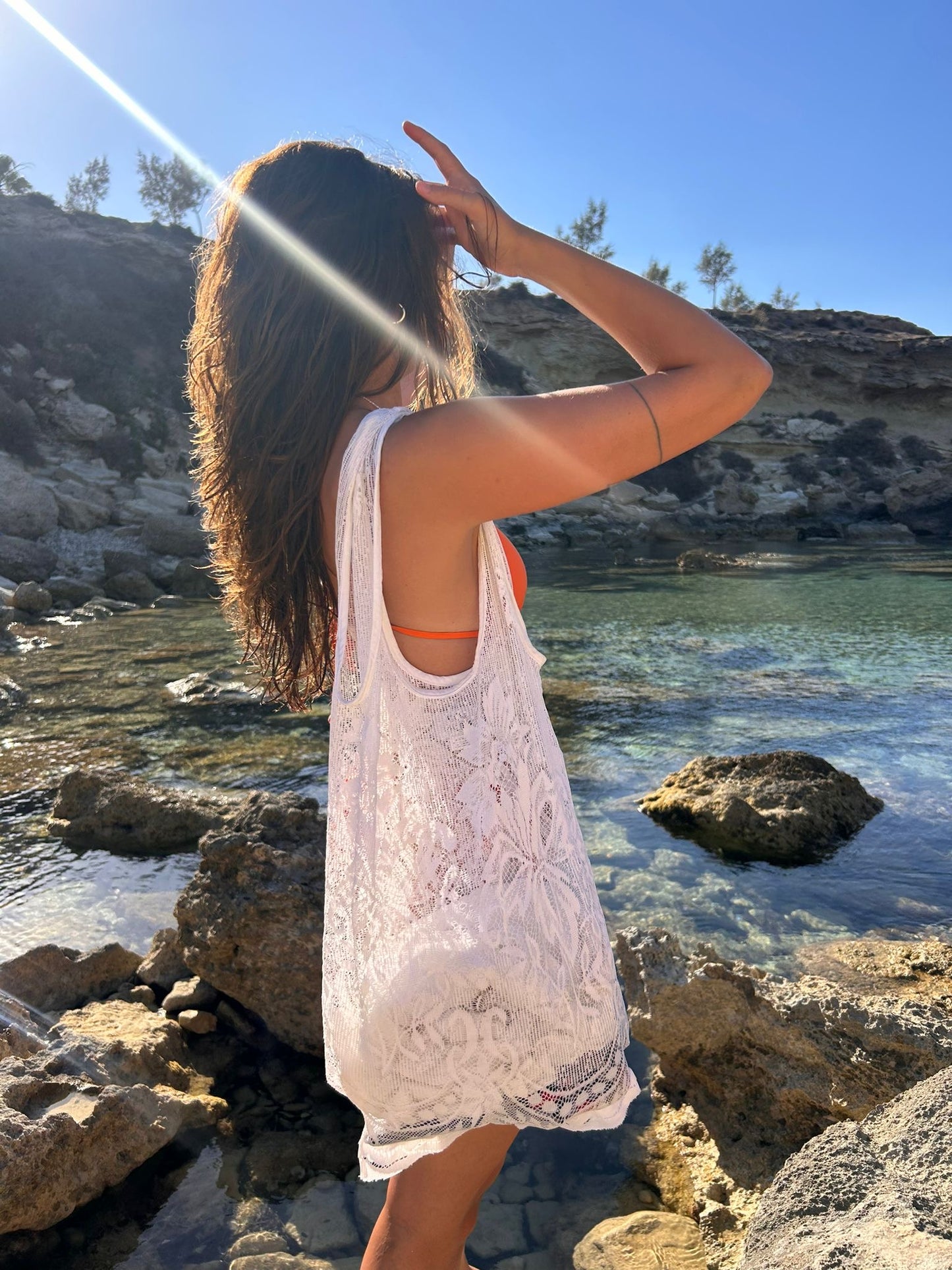 A woman standing on a rocky beach with clear water and blue sky in the background, wearing a white lace tote bag and orange bikini top.