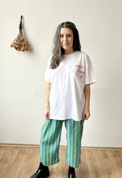 Model wearing a white t-shirt with 'Meine Liebe' embroidery, paired with striped green pants, showcasing a classic look.