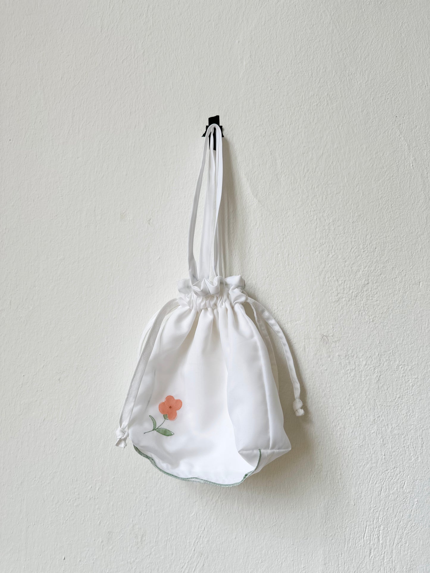 A white mini tote bag with a floral applique on the front, hanging against a plain wall.