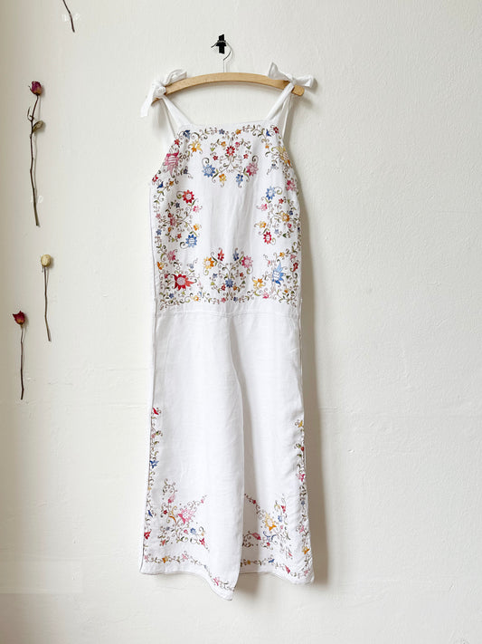 White overalls/jumpsuit with colourful floral embroidery hangs on a wall.