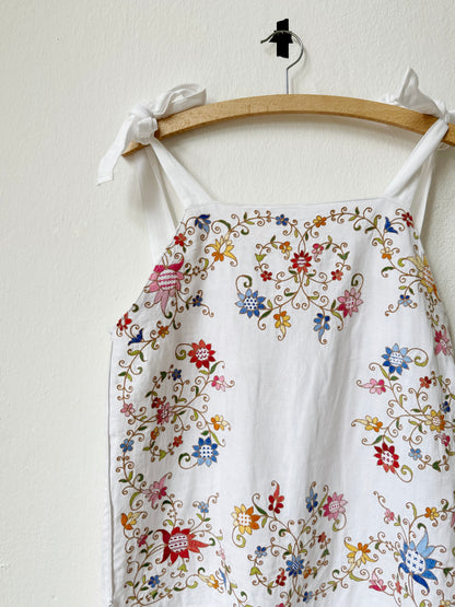White jumpsuit with colourful floral embroidery and tie straps hangs on a wall.